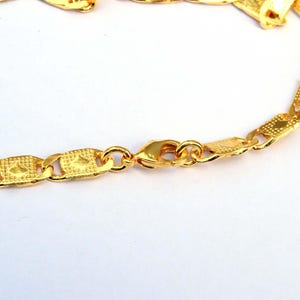 May include: Close-up of a gold-colored chain necklace with a lobster clasp. The chain links have a textured, geometric design. The necklace is on a white background.