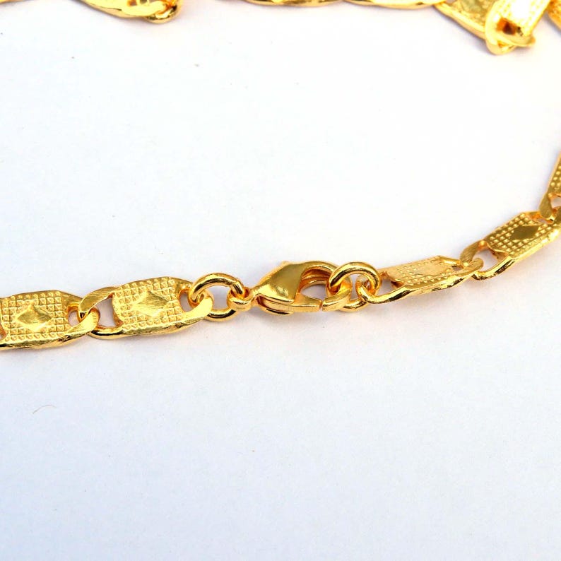May include: Close-up of a gold-colored chain necklace with a lobster clasp. The chain links have a textured, geometric design. The necklace is on a white background.