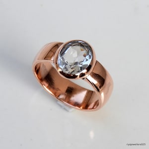 Op de afbeelding: Een roségouden ring met een grote, ovaal geslepen, heldere edelsteen. De band is breed en gepolijst, met een glad, afgerond ontwerp. De edelsteen is gezet in een bezel setting. De ring ligt op een wit oppervlak.