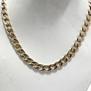 Op de afbeelding: Een goudkleurige gourmetketting. De ketting wordt gepresenteerd op een wit oppervlak. De schakels van de ketting zijn dik en nauw met elkaar verbonden, wat een gedurfde en klassieke uitstraling creëert. Het metaal heeft een gepolijste afwerking, die licht reflecteert.