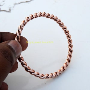 May include: A copper-colored, twisted metal bangle bracelet. The bracelet has a rope-like design and is held by a hand against a white background. The jewelry is circular and appears to be made of a solid metal.