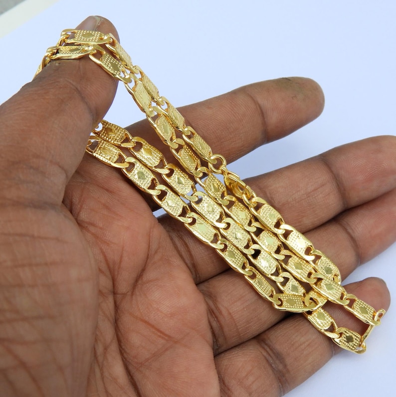 May include: A gold-colored chain necklace with a repeating rectangular link pattern. The chain is held in a hand against a white background. The links have a textured design.