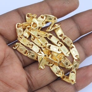 May include: A gold-colored chain necklace with a unique link design. The links are rectangular with a textured pattern and diamond-shaped accents. The necklace is displayed on a hand.