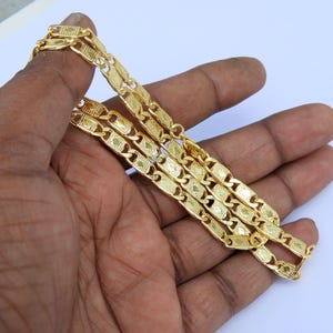 May include: A gold-colored chain necklace with a repeating rectangular link design. The chain is held in a hand against a white background. The links have a textured pattern.