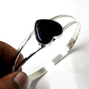 May include: A silver bangle bracelet with a black heart-shaped gemstone. The bracelet is open and has a simple design.
