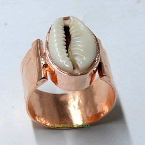 May include: A copper-colored ring featuring a large, off-white cowrie shell. The ring has a wide band with a hammered texture and a rectangular accent on each side of the shell setting. The shell is oval-shaped and has a natural, ridged texture.