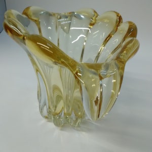 May include: A clear glass bowl with a yellow tint and a wavy, organic shape. The bowl has a wide, shallow opening and a rounded base.