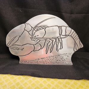 May include: A silver-toned metal napkin holder featuring a cutout silhouette of a lobster. The lobster design is etched with dark lines, and the base of the holder has a textured, speckled finish that mimics a sandy or pebbled ocean floor.