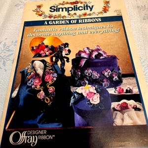 May include: A vintage Simplicity pattern cover titled "A Garden of Ribbons." The cover features a dark blue vest and two hats decorated with ribbon flowers. The text reads "Fantastic ribbon techniques to decorate anything and everything!"