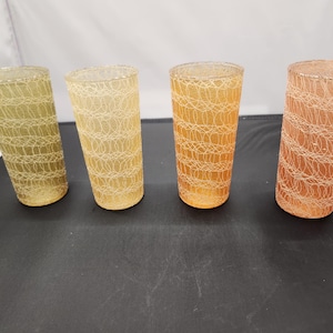 May include: Four tall, cylindrical drinking glasses in shades of yellow, orange, and peach. Each glass features a delicate, white, web-like pattern. The glasses are arranged in a row, showcasing their varying colors and design.