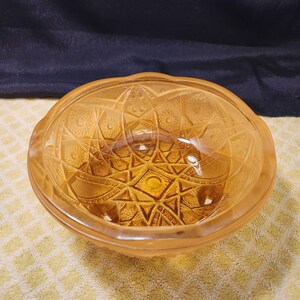 May include: A round amber-colored glass bowl featuring a pressed geometric pattern with starbursts, diamonds, and circular motifs. The bowl has a scalloped rim and a thick, transparent construction that catches light, highlighting the intricate relief.