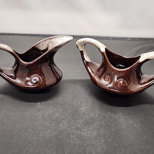 May include: Two brown ceramic gravy boats with a unique design. Each boat has a handle and a spout, with a white and brown speckled rim. The boats have a swirl design on the front.