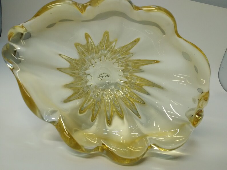 May include: A yellow glass bowl with a starburst design on the bottom. The bowl is made in Italy and has the words "Made in Italy" and "Venini Murano" on the bottom.