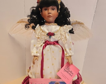 Może przedstawiać: Porcelanowa lalka z ciemnobrązowymi włosami i białą sukienką w złote gwiazdki i czerwoną wstążkę. Lalka ma skrzydła anioła i nosi czerwoną i złotą koronę. Lalka trzyma etykietę z napisem "Paradise Galleries".