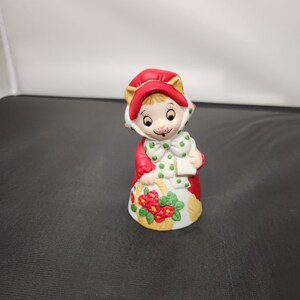 May include: A ceramic figurine of a character wearing a red hooded cloak and a white dress with a floral basket design. The character has a white bow with green polka dots and a smiling face. The figurine is a decorative item.
