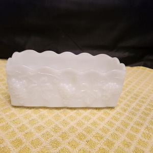 May include: A rectangular, white glass dish with a scalloped rim. The sides are decorated with a grape and leaf pattern. The dish is sitting on a yellow patterned surface.