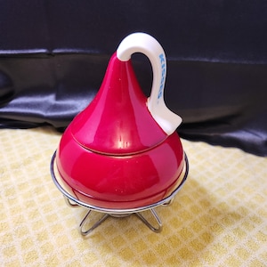 May include: A red ceramic candy dish shaped like a Hershey's Kiss, with a white handle and the word "KISSES" printed on it. The dish sits on a silver metal stand with three legs. The background is a yellow patterned cloth.