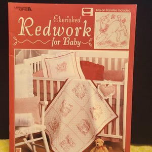 May include: A red and white baby bedding set with a matching quilt, pillow, and crib bumper. The quilt features embroidered red designs on a white background. The set is displayed in a crib, with a teddy bear in the foreground. The book cover reads "Cherished Redwork for Baby."
