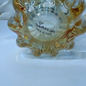 May include: A clear glass seashell-shaped decorative object with a gold tint. The bottom of the object has a label that reads "Teleflora gift. Made in China."