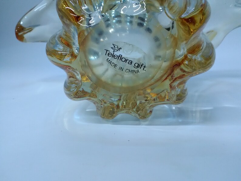 May include: A clear glass seashell-shaped decorative object with a gold tint. The bottom of the object has a label that reads "Teleflora gift. Made in China."
