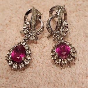 May include: A pair of silver-toned drop earrings. Each earring features a teardrop-shaped, magenta-colored stone surrounded by small, clear stones. The top portion has a looping design with additional clear stones.