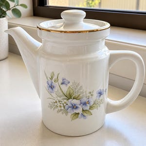 May include: A white ceramic teapot featuring a floral pattern of blue pansies and green leaves on the front. The teapot has a matching lid with a gold-trimmed edge, a curved handle, and a pointed spout. The design is centred on the body.