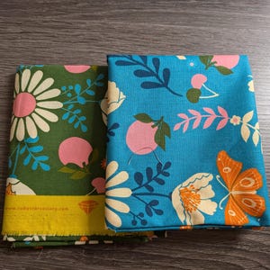 Puede incluir: Dos piezas de tela dobladas con estampados florales. Una es verde con margaritas blancas y detalles rosas, la otra es azul con flores blancas, mariposas naranjas y fruta rosa. La dirección del sitio web www.rubysforsociety.com es visible.