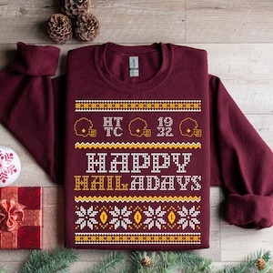 May include: Burgundy sweatshirt with a gold and white holiday pattern. The pattern includes snowflakes, footballs, and the text "HAPPY HOLIDAYS".