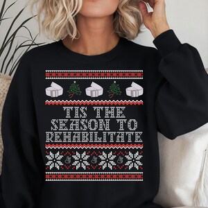 May include: Black sweatshirt with a festive design. The design includes white and red patterns, Christmas trees, and the text "Tis the Season to Rehabilitate" in white. The sweatshirt has long sleeves.