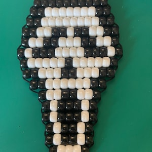 May include: A keychain crafted from black and white plastic pony beads arranged in a geometric pattern that resembles a skull shape. The beads are strung together on a dark cord, and a silver-toned metal lobster clasp is attached to the top.