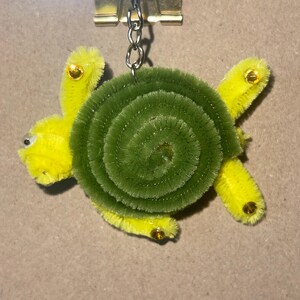 May include: A keychain featuring a turtle shape crafted from yellow and green fuzzy pipe cleaners. The turtle has a coiled green shell, yellow limbs, and small gold-colored circular accents on its legs. A silver metal chain and ring are attached.