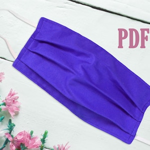 May include: A purple fabric face mask with white elastic ear loops. The mask has pleats and is lying on a white wooden surface. The text "PDF" is in pink letters in the upper right corner of the image.