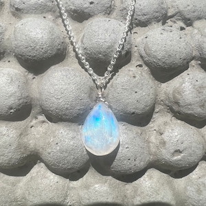 May include: A silver chain necklace with a pear-shaped, iridescent moonstone pendant. The moonstone displays a play of colors, including blue and white. The pendant is wire-wrapped and suspended from a delicate chain.
