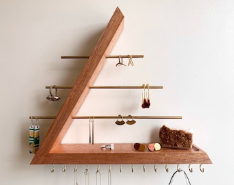 May include: A triangular wooden jewelry organizer with brass rods and hooks. The organizer has a shelf for rings and other small items. There are several pairs of earrings hanging from the rods, and a small brown pouch is sitting on the shelf.