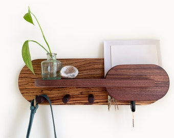 May include: A wooden wall-mounted key holder with a shelf, three hooks, and a small oval-shaped compartment. The key holder is made of dark wood with a natural wood grain. A small glass vase with a green plant is on the shelf, and a small white crystal is next to it. A teal leather strap hangs from one of the hooks.
