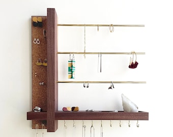May include: A wooden jewelry organizer with a dark brown finish, featuring brass rods and a corkboard backing. Various earrings and necklaces are displayed. A small shelf holds additional jewelry and a decorative crystal. Hooks are along the bottom.