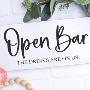 Puede incluir: Un letrero de madera blanco con caligrafía negra que dice "Open Bar The Drinks Are On Us!"