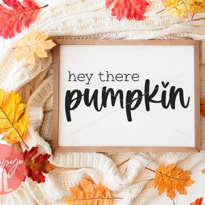 Peut inclure: Une pancarte blanche avec un cadre en bois, posée sur une couverture en tricot blanc avec des feuilles d'automne orange et rouge. La pancarte dit "hey there pumpkin" en écriture noire.