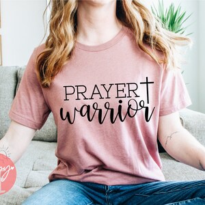 Puede incluir: Una camiseta rosa con el texto "PRAYER warrior" y una cruz impresa.