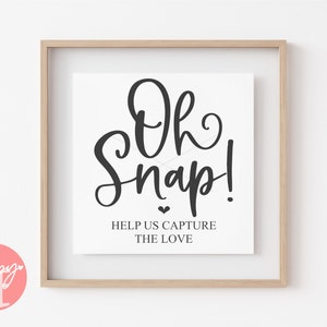 May include: A white sign with black text that reads "Oh Snap! Help us capture the love." The sign is framed in a light wood frame.