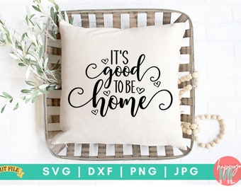 May include: A white throw pillow with black text that reads "It's good to be home" with small hearts surrounding the text. The pillow is sitting in a wooden tray with a white background.