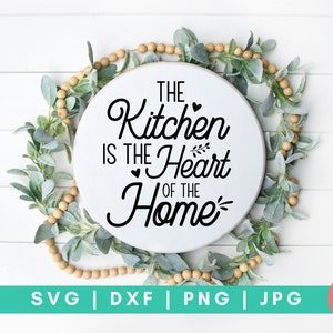 May include: A white round sign with black text that reads "The Kitchen is the Heart of the Home". The sign is surrounded by a garland of green leaves and wooden beads.