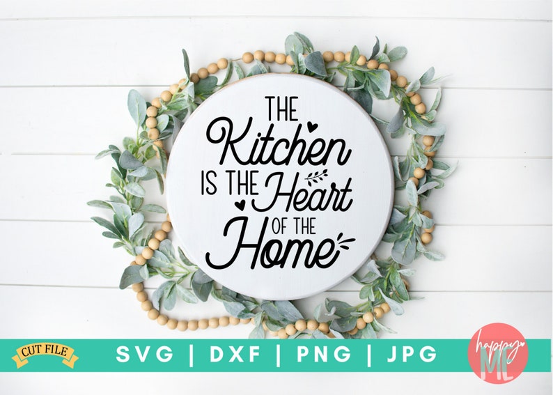 May include: A white round sign with black text that reads "The Kitchen is the Heart of the Home". The sign is surrounded by a garland of green leaves and wooden beads.
