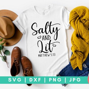 以下が含まれることがあります： 白いTシャツに、黒い文字で「Salty and Lit」と書いてあり、「and」の両側にハートマークがあります。テキストの下には「Matthew 5:13」と書いてあります。