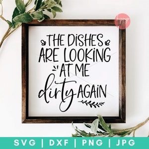 May include: A white sign with a brown wooden frame, featuring the text "THE DISHES ARE LOOKING AT ME dirty AGAIN" in black cursive font. The sign is decorated with a small sprig of leaves.