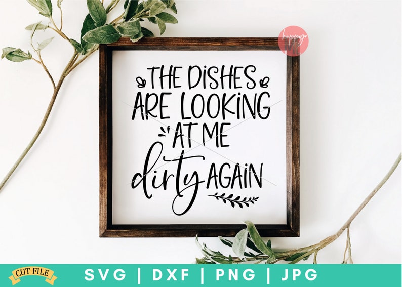 May include: A white sign with a brown wooden frame, featuring the text "THE DISHES ARE LOOKING AT ME dirty AGAIN" in black cursive font. The sign is decorated with a small sprig of leaves.