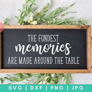 May include: A wooden sign with a black background and white text that reads "The Fondest Memories Are Made Around The Table". The sign is surrounded by green leaves and white flowers.