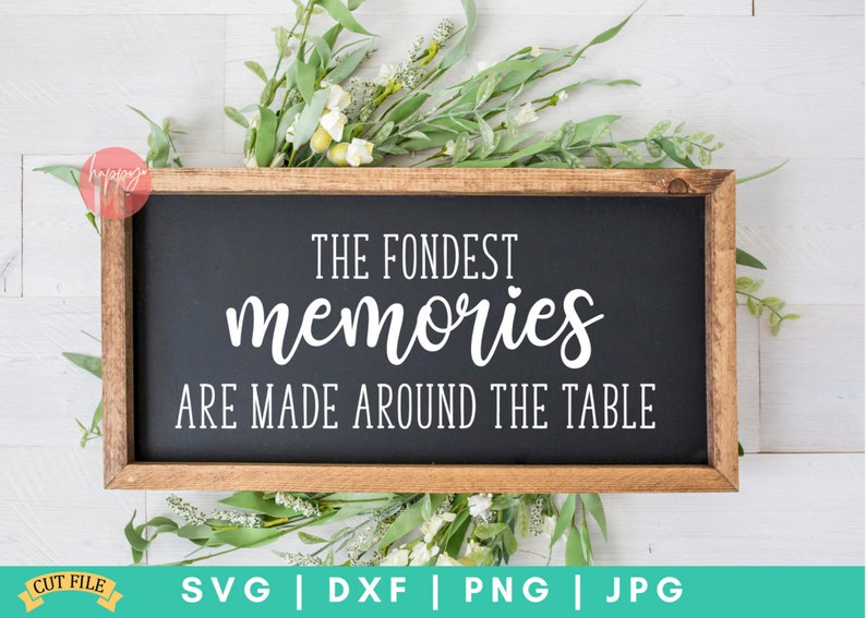 May include: A wooden sign with a black background and white text that reads "The Fondest Memories Are Made Around The Table". The sign is surrounded by green leaves and white flowers.