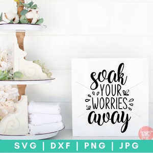 K&ouml;nnte beinhalten: Ein wei&szlig;es Holzschild mit schwarzem Kalligraphietext, der "soak your worries away" lautet. Das Schild steht auf einer wei&szlig;en Oberfl&auml;che mit einem mehrst&ouml;ckigen Tablett mit wei&szlig;en Blumen und Handt&uuml;chern im Hintergrund.