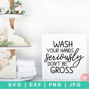 May include: White sign with black text that reads "Wash your hands seriously don't be gross". The sign is sitting on a white shelf with a tiered tray behind it. The tray is decorated with white flowers and white towels.
